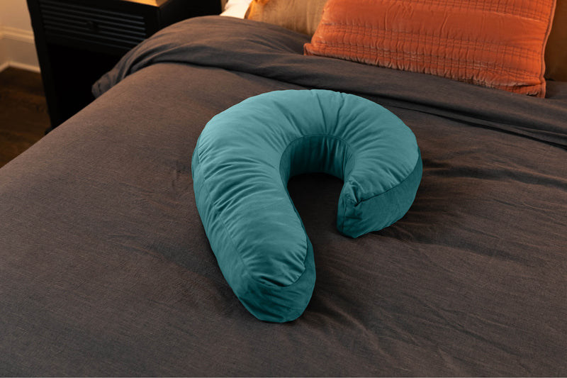 The Liberator Lune Snuggle Sex Toy Mount in Teal sits out on a bed. It looks bigger than the bed pillows at the top of the bed. It looks like one-half of a heart shape. | Kinkly Shop