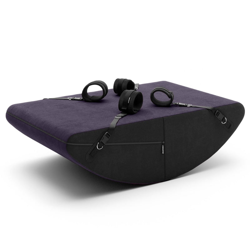 The Liberator Scoop Rocker in Aubergine with all of the included accessories (aside from the blindfold!) sitting on top of the Scoop. Each one of the cuffs is clipped into the Velcro tab connectors which are placed along the four corners of the Scoop Rocker. There's a zipper that runs the underside on half of the shape, outside of where your bare skin would touch it. The material looks soft. | Kinkly Shop