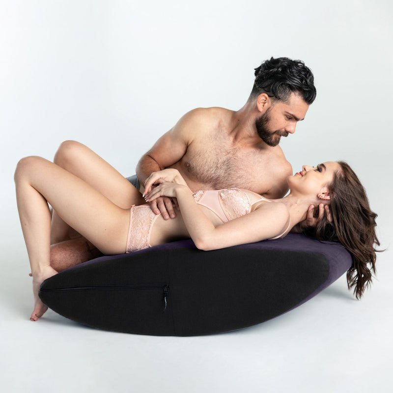 One partner is laying on their back on top of the Liberator Scoop Rocker. Their partner is laying on their side, snuggled up to the first person's side, to look into their eyes for intimacy. Both partners are allowing the Liberator Scoop Rocker to lean towards their feet, lifting up the laying partner's face for easier smooching. | Kinkly Shop