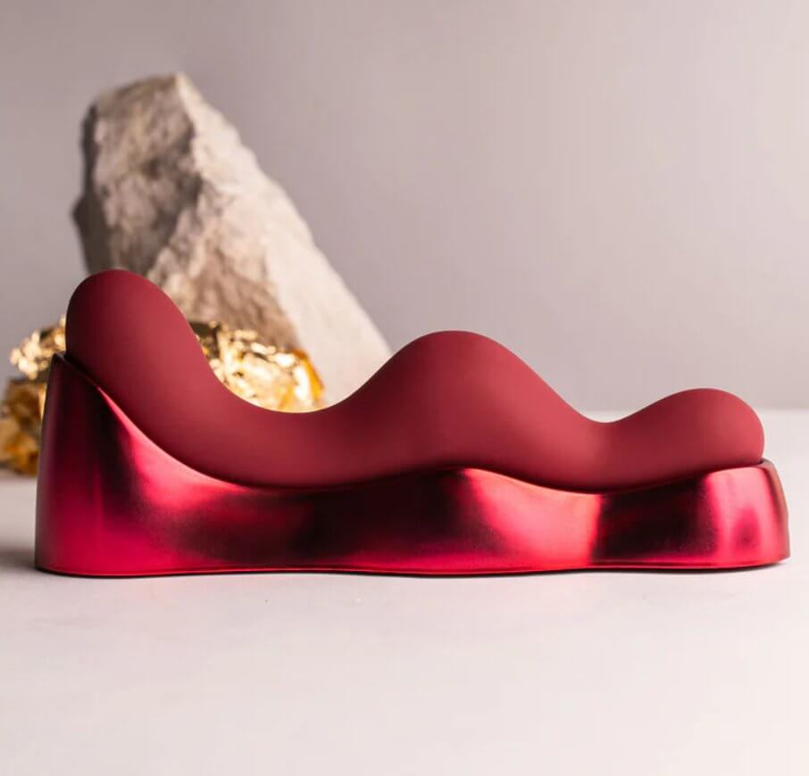 The Rocks-Off Ruby Glow Blush rests in its cradle with a side profile angle. This showcases the serious curves along the length of the vibrator. The tallest end is very tall and designed to nestle between the labia for clitoral vibrations during sit-on pleasure. The middle curve is almost as tall in height, non-penetrating but designed to rest against the sensitive vaginal entrance. There's a small curve at the end of the toy, designed for non-penetrating sensations of the anal entrance. | KInkly Shop