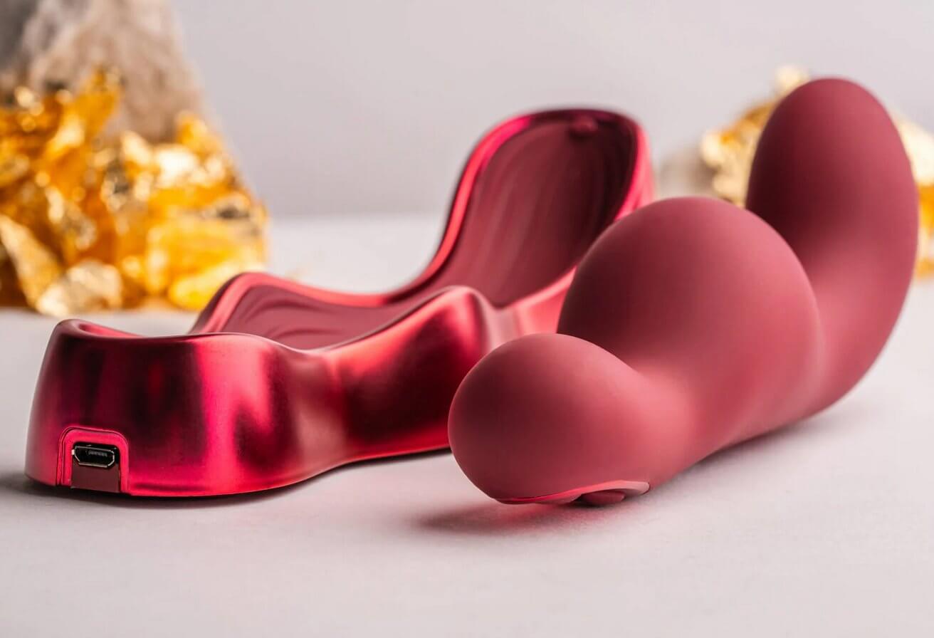 The Rocks-Off Ruby Glow Blush in front of a white background with yellow flowers in the background. The cradle for the vibrator and the vibrator itself are sitting next to one another. The plug-in charging port at the base of the charging cradle can be viewed. | Kinkly Shop