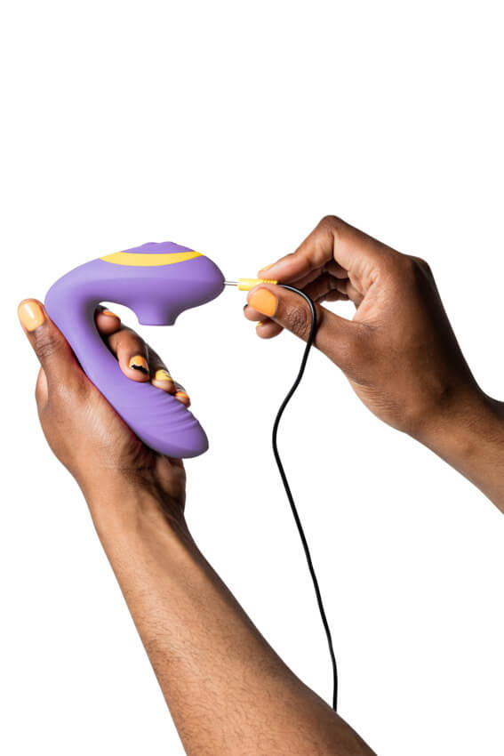 A person holds the ROMP Reverb in their hand. In their opposite hand, they hold the charger for the ROMP Reverb. They are sliding the charger into the tip of the clitoral end to charge the toy. | Kinkly Shop