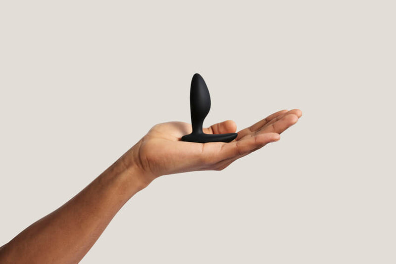 A person's hand is laid flat in front of a white background. On top of it, a We-Vibe Ditto+ is balanced, sitting upright. The plug looks very slender and small in their hands. It looks like it's slightly less thick than two of the person's fingers. | KInkly Shop