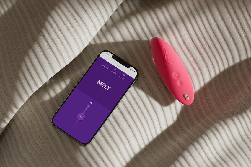 The We-Vibe Melt sitting on top of a comforter next to a cell phone. The cell phone is open to the We-Vibe Melt control panel, showing the slider bar that can adjust the intensity up and down with a drag of a finger. | Kinkly Shop