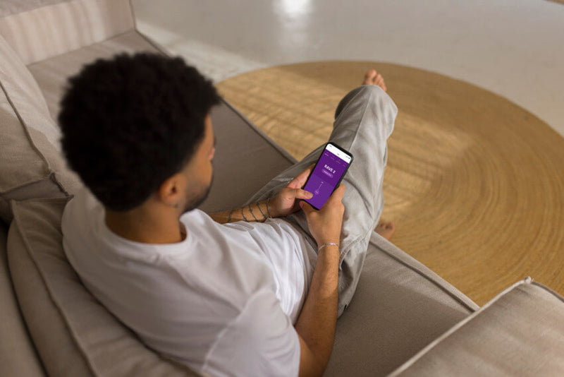 A person sits on their cell phone on the couch. The camera looks over their shoulder. The We-Vibe Rave 2's app control is on the screen of their phone, and they're presumably controlling their partner's vibrator from afar since their partner is nowhere to be seen. | Kinkly Shop