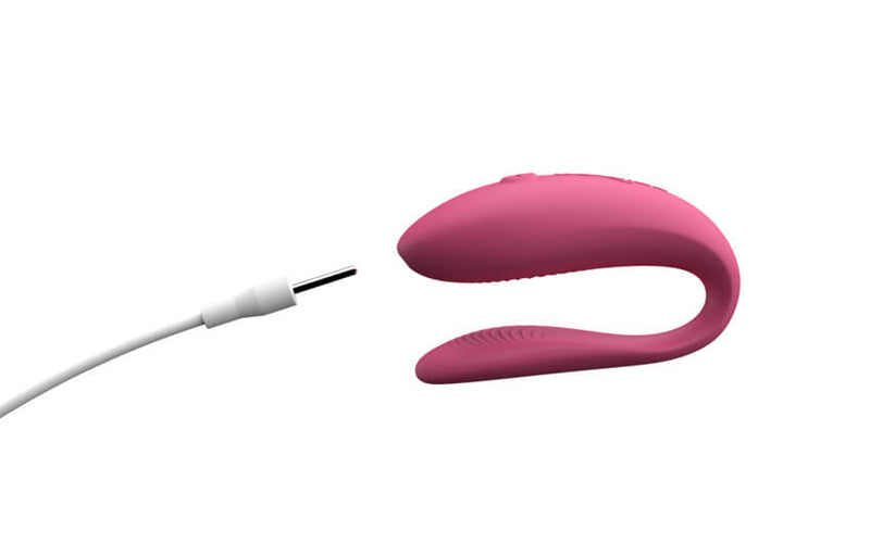 The We-Vibe Sync Lite in Pink up against a white background. The charging cord pin is being pointed towards the tip of the thicker end of the vibrator. This is where the charging port is located. | Kinkly Shop