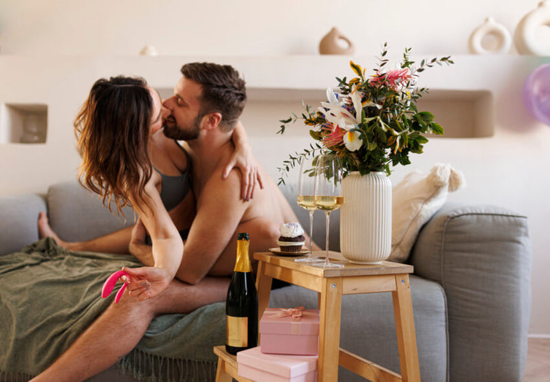 A couple in their underwear embraces on the couch. There is champagne in the table on the foreground. One of them is holding a pink We-Vibe Sync Lite in their hands. It's smaller than the palm of their hand. | Kinkly Shop