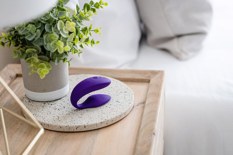 The We-Vibe Sync 2 in Purple sitting on a decorative marbled platform next to a potted plant. There's a bed with white bedding in the background. The image is very bright and airy. | Kinkly Shop