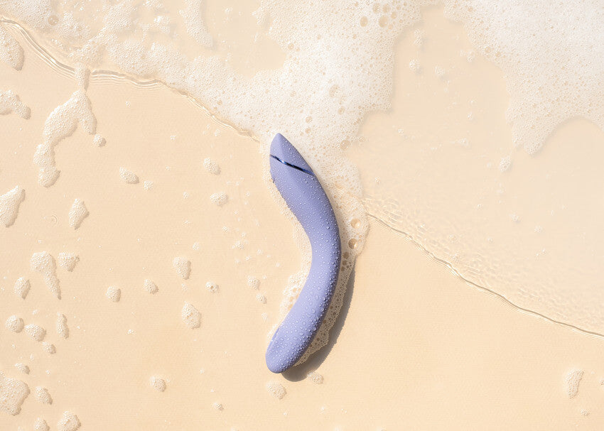 The Womanizer OG laying out on a sand-like surface with a rush of bubbly water reaching out to touch the toy. This showcases its waterproof capabilities. | Kinkly Shop