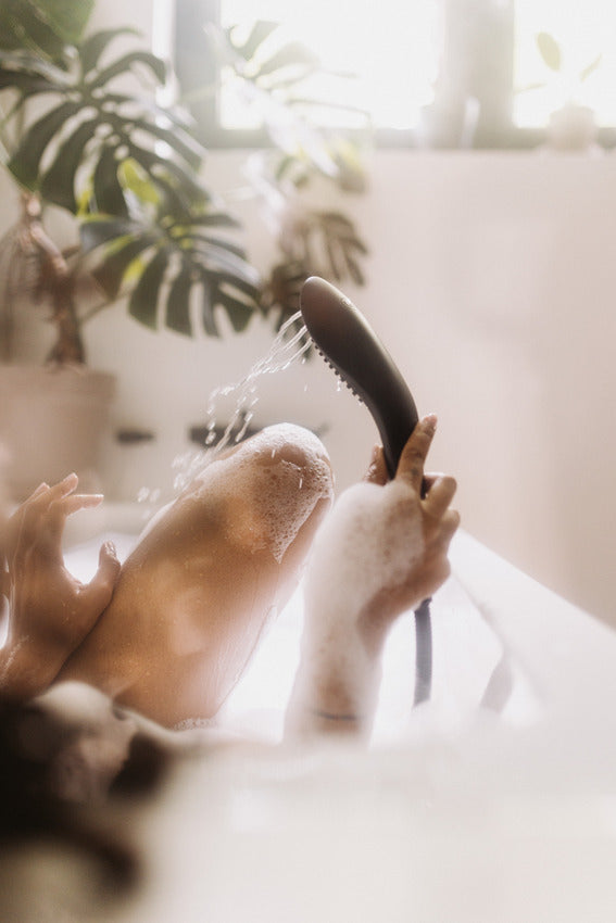 A hazy, relaxing image of a person in their bathtub, using the Womanizer Wave. They have bubbles all over their bare body, and they're comfortably holding the Womanizer Wave while it sprays water onto their bare leg. | Kinkly Shop
