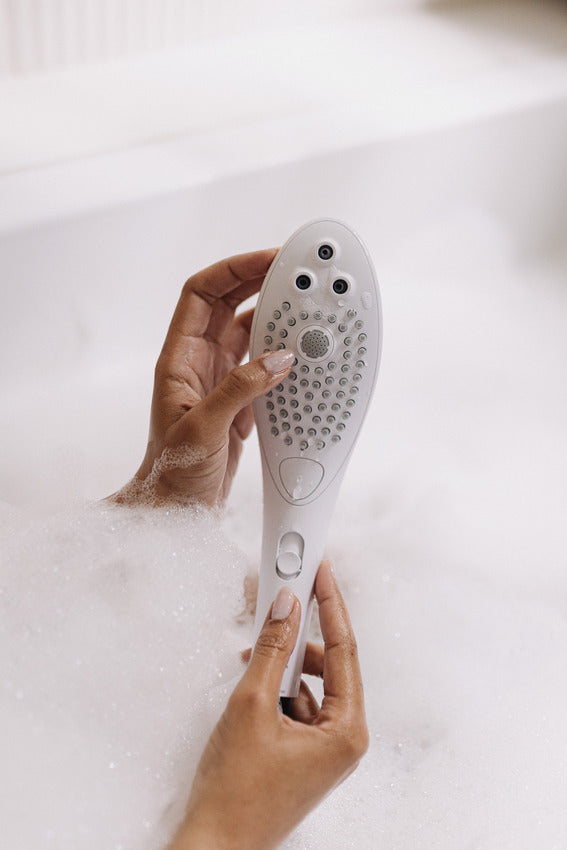 Two hands hold the Womanizer Wave right above the bathtub water that's very bubbly. They're showcasing the size of the showerhead which looks slimmer than most showerheads and slightly larger than most Womanizer sex toys. | Kinkly Shop