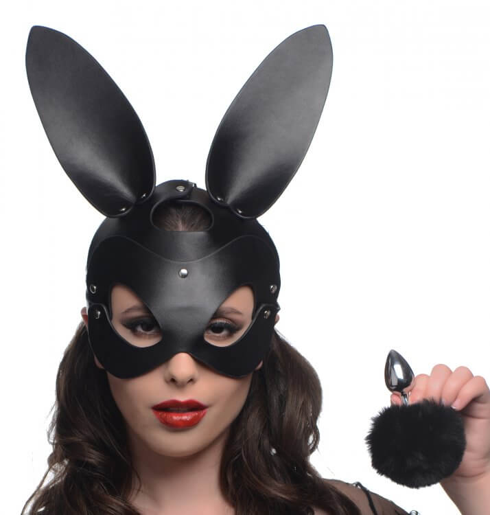 A person is looking into the camera while wearing the bunny mask of the Tailz Bunny Pet Play Kit. In their hand, they are holding the butt plug with the bunny tail attached. It looks like a slim butt plug - no thicker than two of the person's fingers. | Kinkly Shop