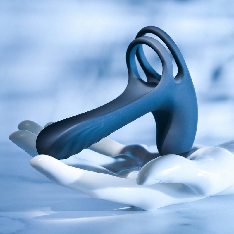 A mannequin hand is laid out flat with the Vibrating Girth Enhancer Extension resting on top of it. The vibrator holds its shape with its rigid silicone, and this angle showcases the two rings at the base of the vibrator that helps anchor it in place as well. | Kinkly Shop