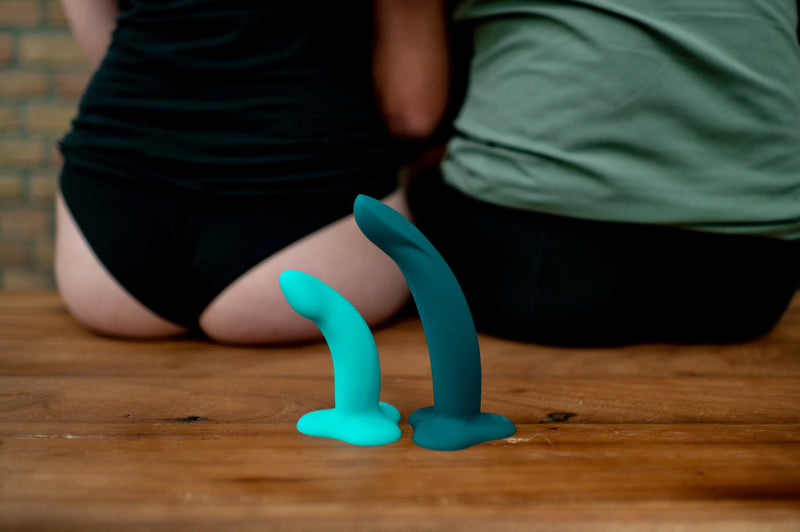 Two people are sitting on a wooden bench. The Limba Flex dildos, in small and medium, are shown sitting upright on their bases behind them. The Medium is clearly longer by a noticeable amount but they look about the same in diameter. | Kinkly Shop