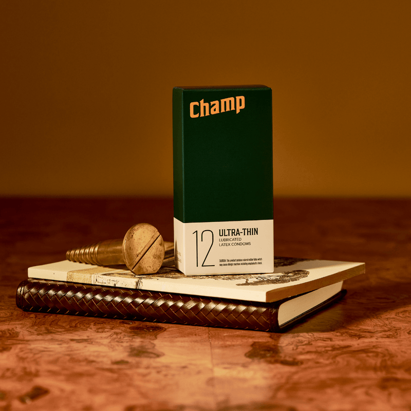 A box of the Champ Ultra-Thin Condoms - 12 Pack condoms sitting on top of a brown journal and a sketchbook. The wall in the background is brown and the stack is resting on top of a brown countertop. The coloration of the image is very orange for a regal-looking photo. | Kinkly Shop