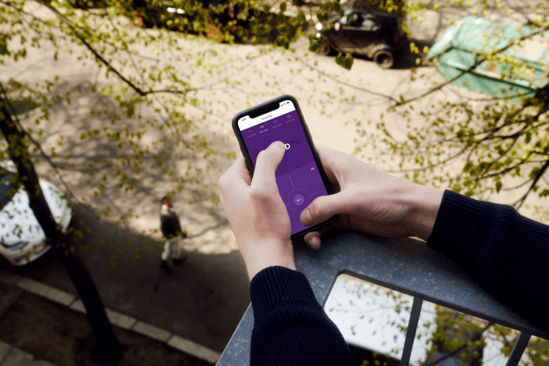 A person holds a cell phone that has the We-Vibe app open to control a vibrator. They are standing on a balcony. Someone else, standing on the street below, looks up at them as the person holding the cell phone increases the vibrations on the app for the We-Vibe Bond. | Kinkly Shop