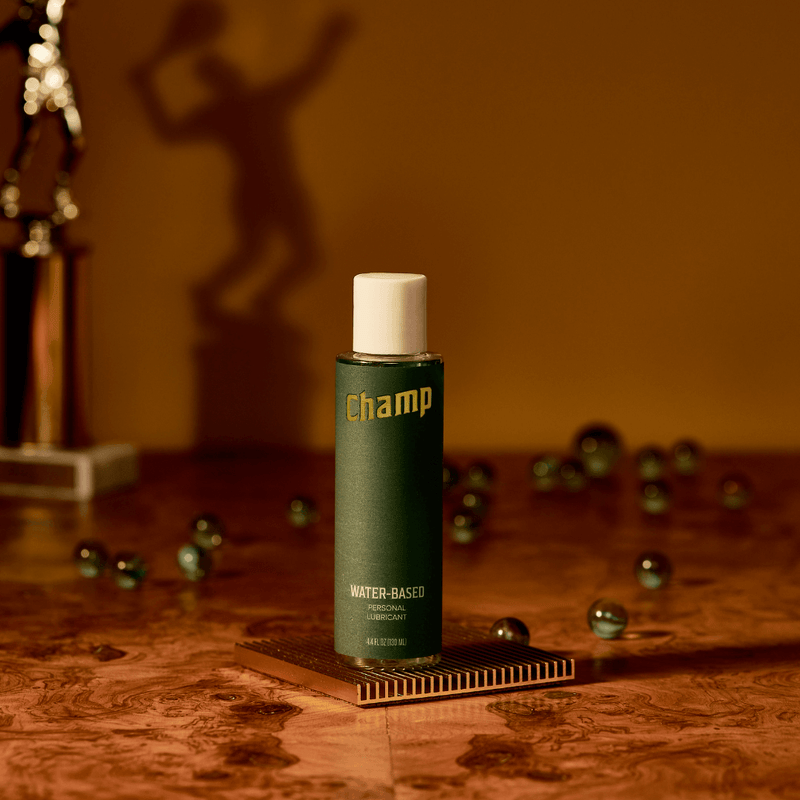 A bottle of the Champ Water-Based Lube - 4.4OZ against a brown wall on top of a brown countertop. There's a sports trophy in the background. The image looks very regal. | Kinkly Shop