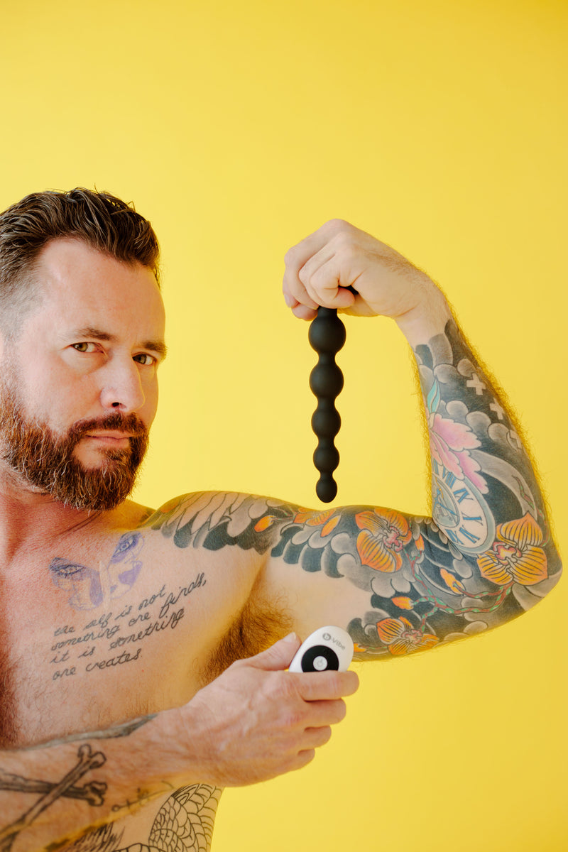 A person with arm tattoos holds up the Cinco anal beads while flexing their bicep. Their other hand holds the remote. | Kinkly Shop