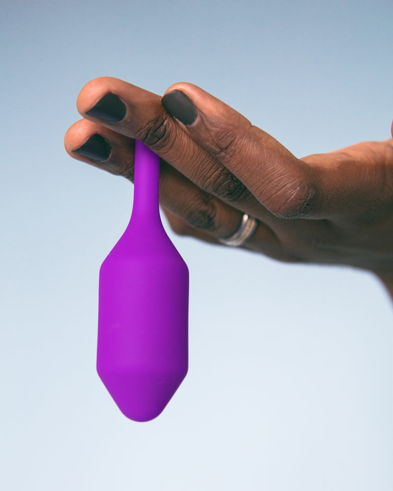 A hand holds the b-Vibe Snug Plug 2 in Purple in-between their fingers. The torpedo, insertable-end hangs down between the fingers. | Kinkly Shop