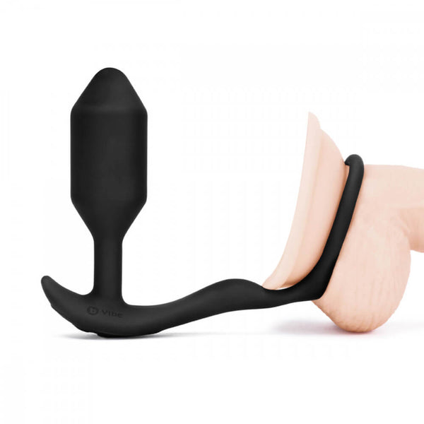 The b-Vibe Vibrating Snug and Tug in Medium against a white background. The plug sits upright while the attached cock ring is wrapped around the base of a dildo. | Kinkly Shop