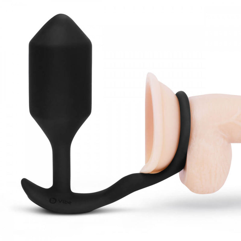 The b-Vibe Vibrating Snug and Tug in Extra-Large against a white background. The plug sits upright while the attached cock ring is wrapped around the base of a dildo. | Kinkly Shop