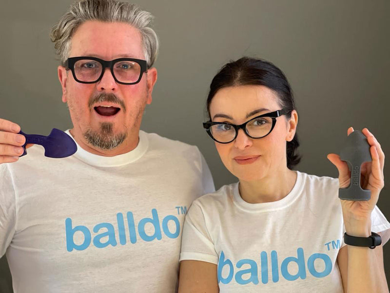 A portrait of the two founders of the Balldo. They're both wearing Balldo t-shirts and holding a Balldo in their hands. Both seem smiling and approachable. This image displays the size of the Balldo compared to a person's hand. The base Balldo with no additional rings is slightly shorter than a smaller hand's length from base of palm to fingertip. | Kinkly Shop