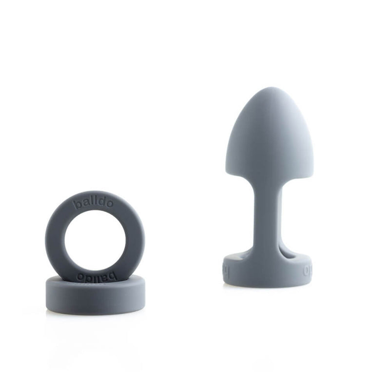The Balldo in Steel Grey. The image shows the Balldo itself sitting upright which is a great angle to show off its extremely tapered tip that looks like a torpedo tip. Next to the Balldo are the two spacer rings. The image shows the word "Balldo" imprinted on the silicone of each of the three pieces. | Kinkly Shop