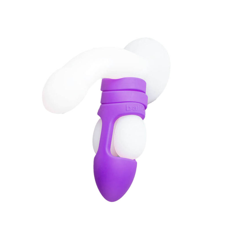 The Balldo in Purple is shown being used on the testicles of a clear dildo. The image shows how the Balldo works - which is by wrapping around the testicles while simultaneously leaving space for the testicles to receive pleasure during penetration. | Kinkly Shop