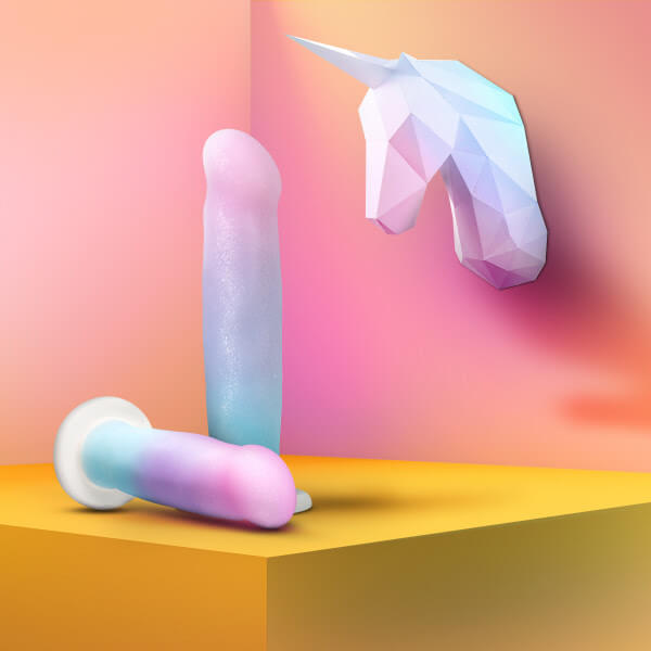 Two Blush Avant D17 Lucky pastel dildos sit on a bright yellow box in front of a peach wall. A unicorn motif in the background matches the exact same shades that the dildos are made from. | Kinkly Shop