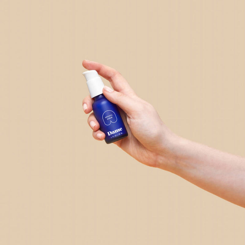 A hand holds the Dame Arousal Serum bottle against a plain brown background. The bottle easily fits into the person's hand. It looks comfortable and easy to pump. | Kinkly Shop