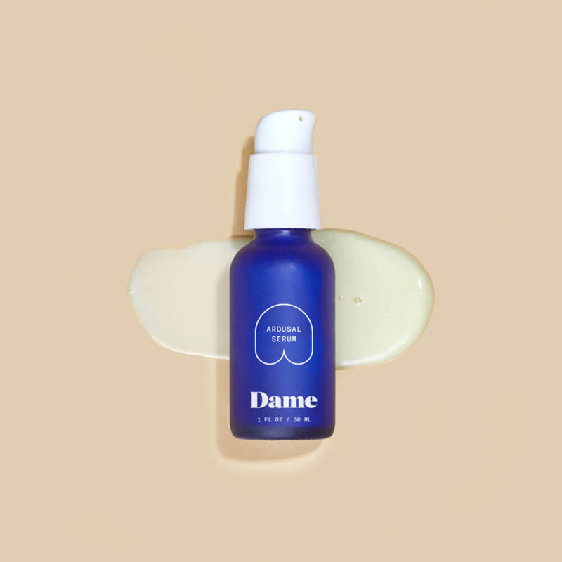 The Dame Arousal Serum lays flat on a brown background. Some of the arousal serum is squirted in a puddle that the blue bottle is laying in. | Kinkly Shop
