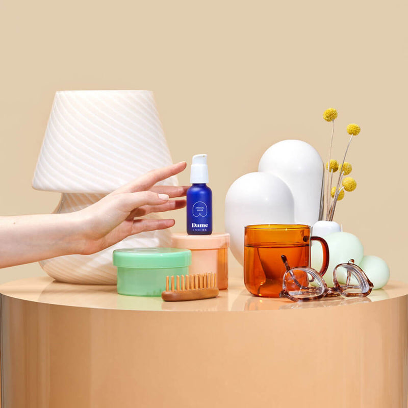 A hand reaches for a Dame Arousal Serum bottle sitting on a bedside table. There are a lot of other bedside accessories next to it including a mug, eyeglasses, a skin brush, and skincare cream tub. | Kinkly Shop