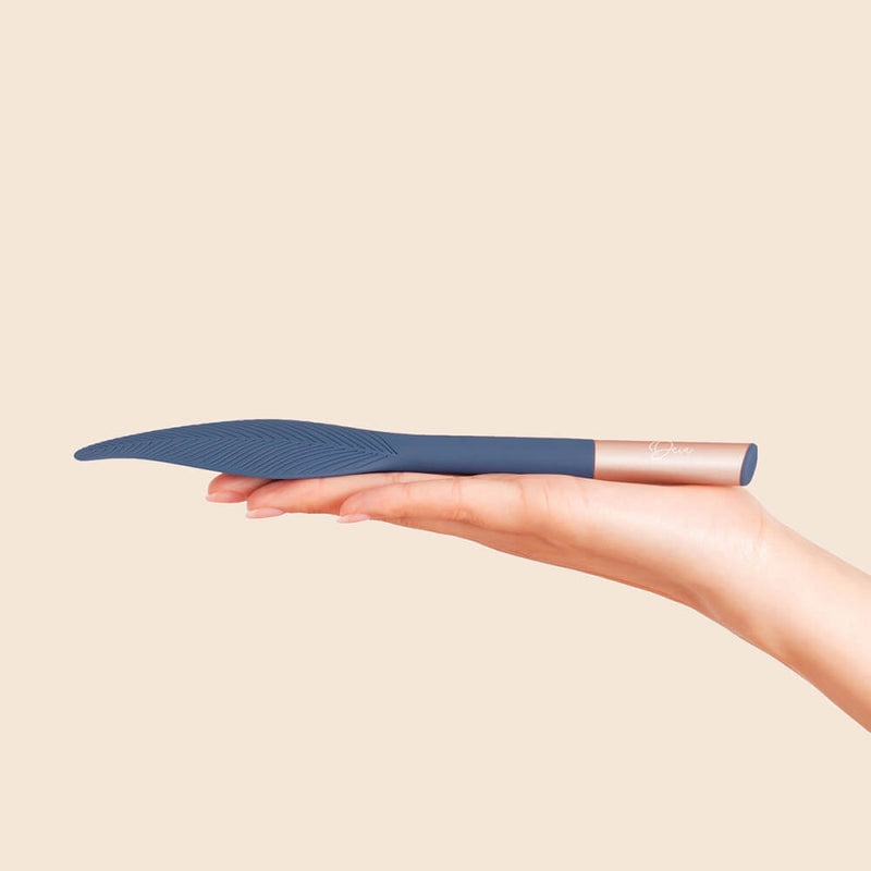 A hand is laid out flat in front of a brown background. The Deia Feather vibrator rests on top of the hand. The vibrator extends past the person's fingertips as it is much longer than the length of an outstretched hand. | Kinkly Shop