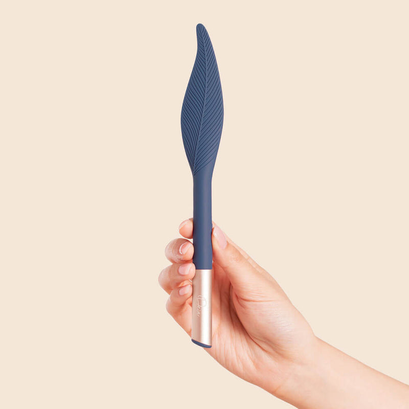 A hand holds the Deia Feather vibrator upright in front of a brown background. The tip of the vibrator is in a "stereotypical" feather shape with a wide, textured base that continues into a slim, soft, rounded tip. The shaft of the vibrator near the handle is untextured. | Kinkly Shop