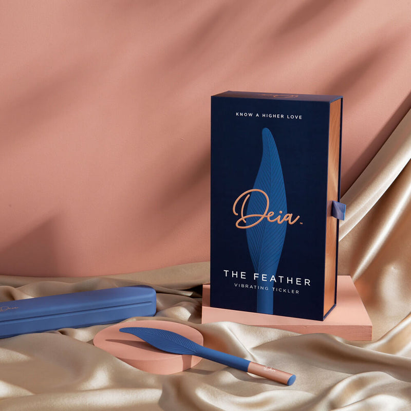 The Deia Feather vibrator lays out on gold satin sheets in front of the packaging for the Deia Feather vibrator. The matching blue storage/charging case is sitting nearby. It looks very sturdy and looks like it's about the size of a pencil case. | Kinkly Shop