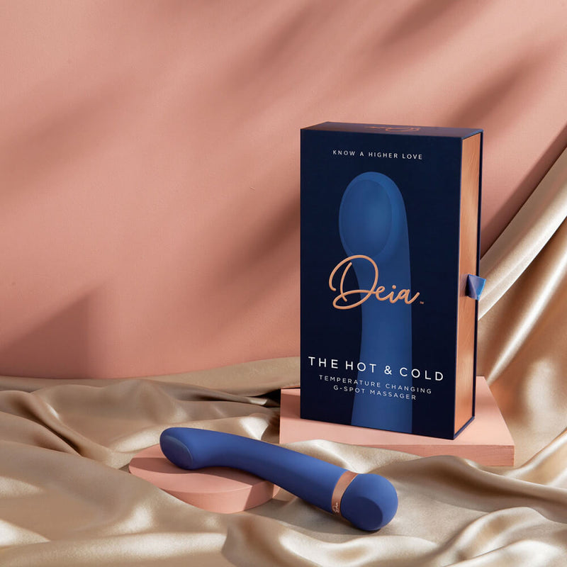 The Deia Hot & Cold Temperature Play Vibrator vibrator rests on a pile of golden silk in front of the box for the vibrator. | Kinkly Shop