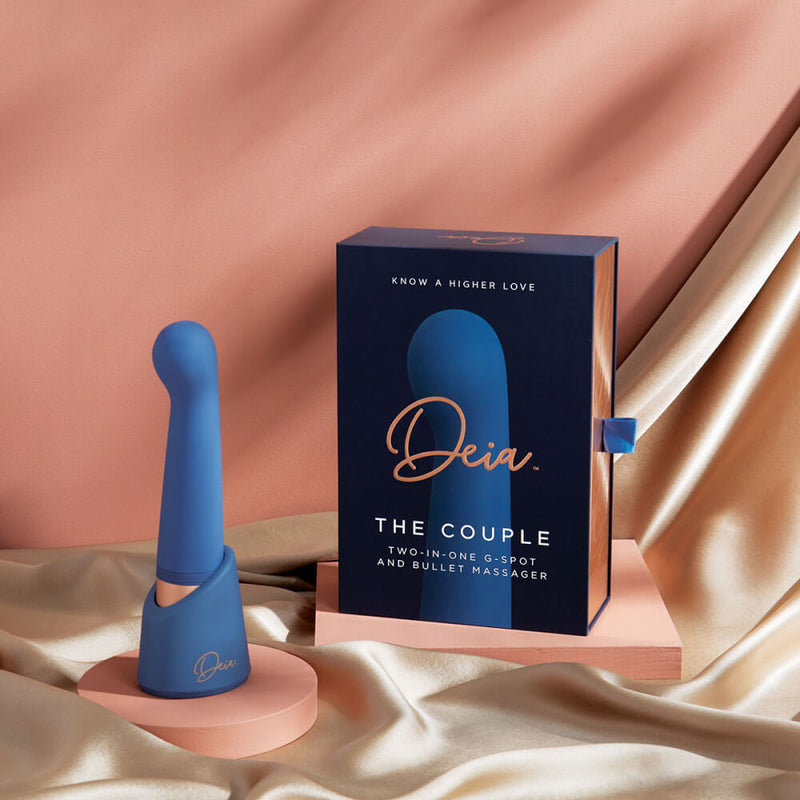 The Deia The Couple interchangeable tip vibrator is sitting upright in its charging/storage base right next to the packaging for the Deia The Couple interchangeable tip vibrator. The base is branded with the "Deia" logo and also color matches the vibrator itself. | Kinkly Shop