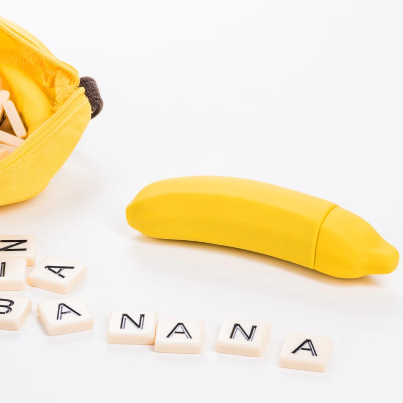 The Emojibator Banana laying out with a plain white background with a bag of word board game tiles spilled nearby. The tiles spell out "Banana" in front of where the Banana vibrator is laying. | Kinkly Shop