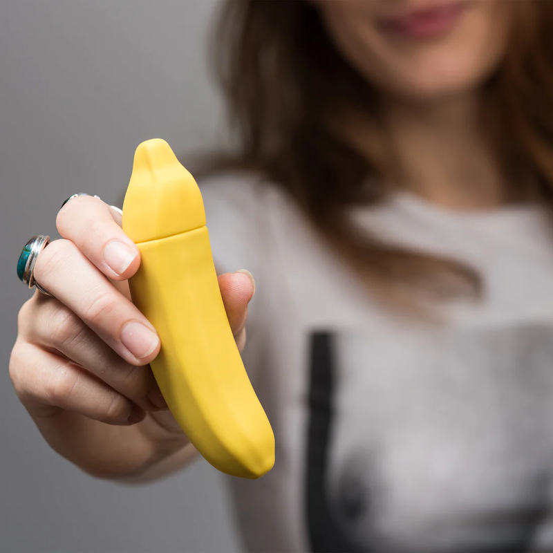 A person holds the Emojibator Banana, pushing it towards the camera. The vibrator looks longer than their fingers but not longer than their entire fingertip-to-palm length. | Kinkly Shop