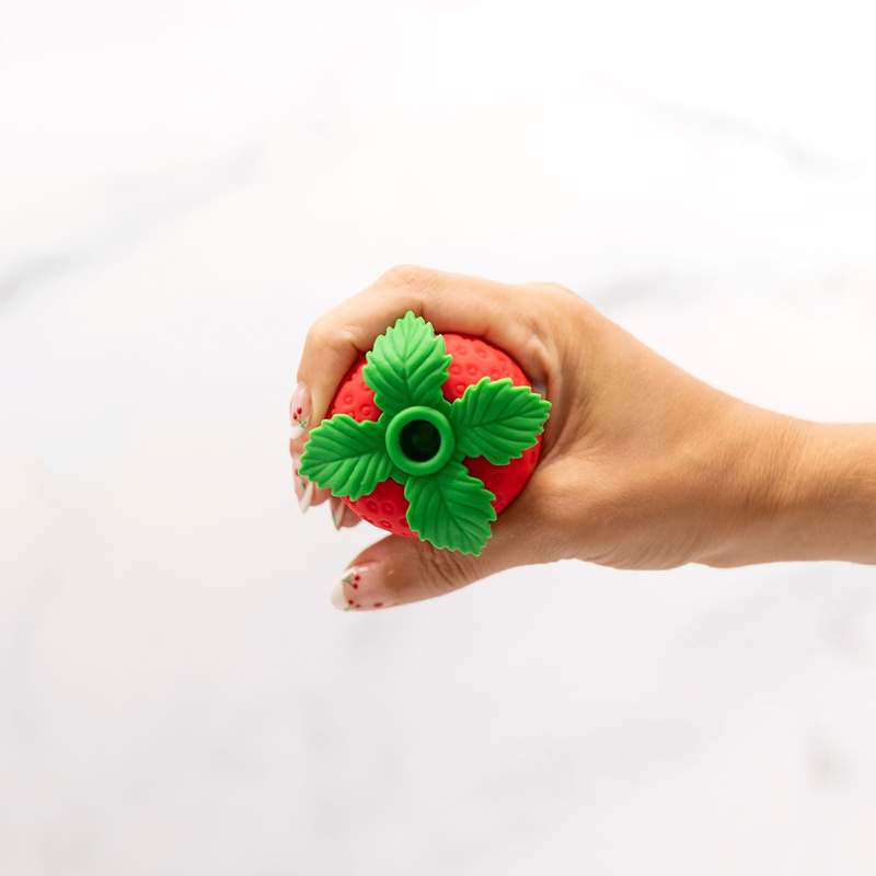 A hand holds the top of the Emojibator Strawberry, with the leaves, close to the camera. This angle showcases the open hole at the tip of the toy that functions as the air suction hole. | Kinkly Shop