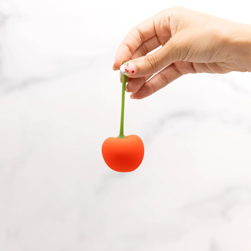 A hand holds the tip of the stem of the Emojibator Cherry. The vibrator itself dangles, looking like an extra-large cherry, from the stem of the vibrator. | Kinkly Shop