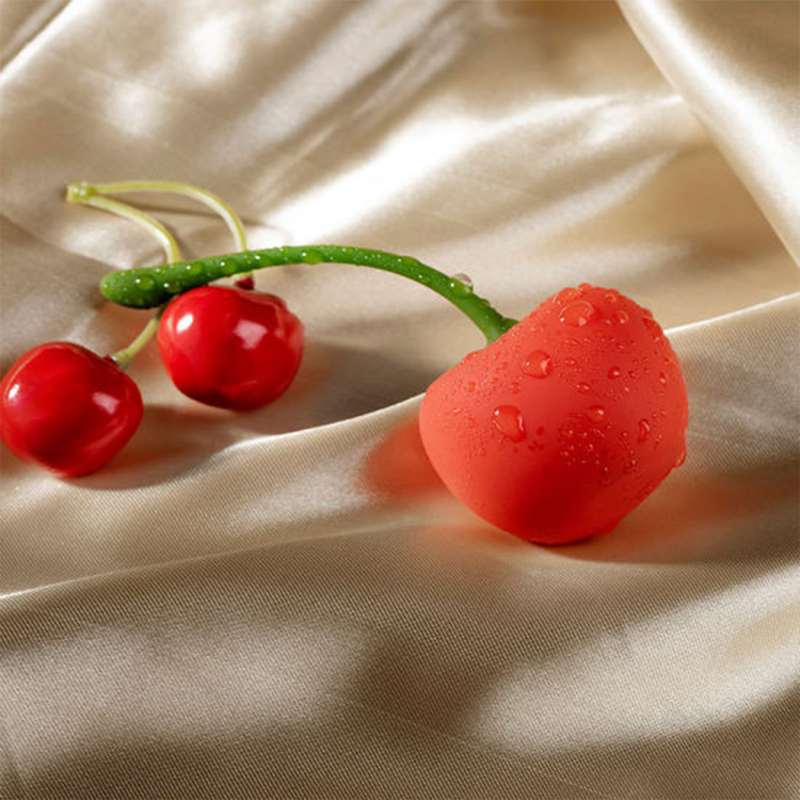 The Emojibator Cherry rests next to two real cherries against peach-colored satin sheets. The Emojibator Cherry is covered in water droplets, showcasing its waterproof capabilities. The Emojibator Cherry is much, much larger than the biological cherries - about 3 times as large. | Kinkly Shop