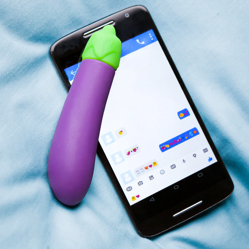 The Emojibator Eggplant sits on top of a cell phone. The cell phone is displaying a back-and-forth text conversation that consists of only emojis. | Kinkly Shop