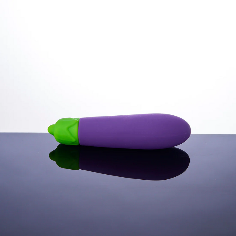 The Emojibator Eggplant laying on a flat surface. The Eggplant does not have any curve to its shaft. The purple is a vibrant purple hue with a bright green that isn't quite lime green. The shaft of the Eggplant has no texture to it, but it's widest at near the tip (away from the green stem) of the toy. | Kinkly Shop