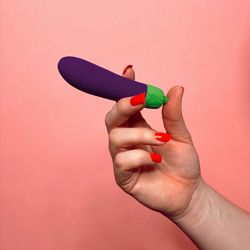 A person's hand holds the Emojibator Eggplant like a tampon. The toy is thicker than a finger but not as thick as two fingers. It looks like it's about an inch longer than the person's longest finger. | Kinkly Shop