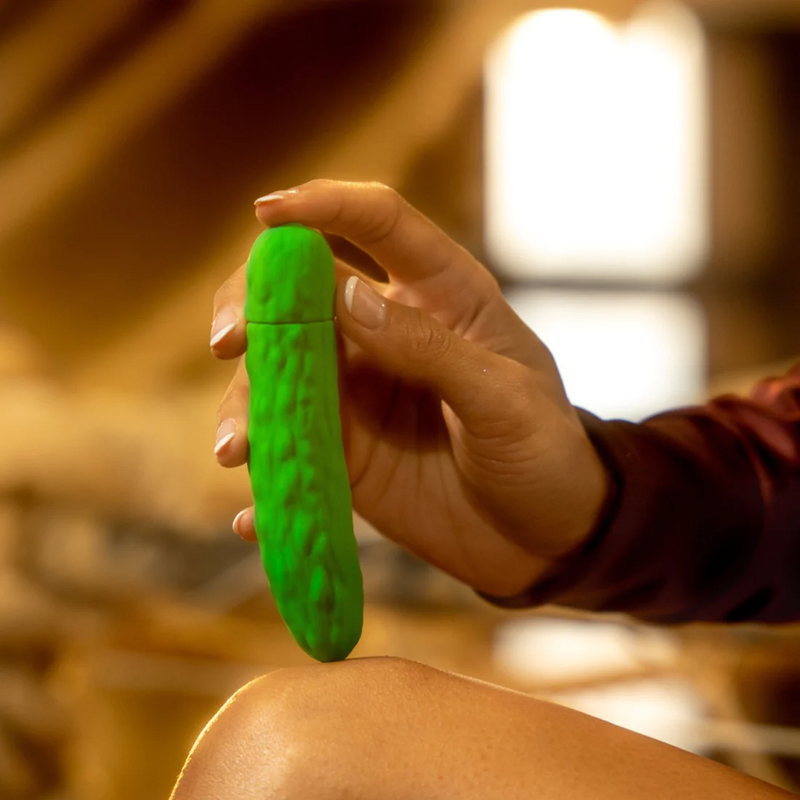 A person holds the Emojibator Pickle in their hand, pressing the end of the vibrator against their knee. This shadowed angle showcases the "pickle" texturing all over the length of the green vibrator. It has a bunch of random lumps and bumps and uneven, smooth texture bumps. | Kinkly Shop