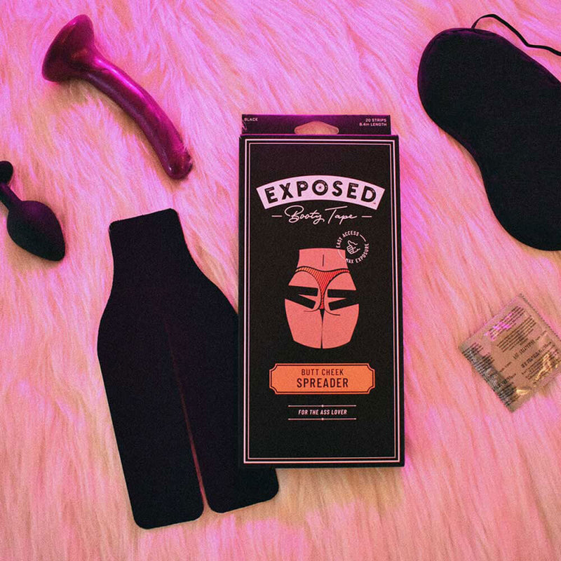 The packaging for the Exposed Booty Tape is shown in center frame. Around it, you can see the Exposed Booty Tape strips, a slender dildo, a small butt plug, a blindfold, and a condom. | Kinkly Shop