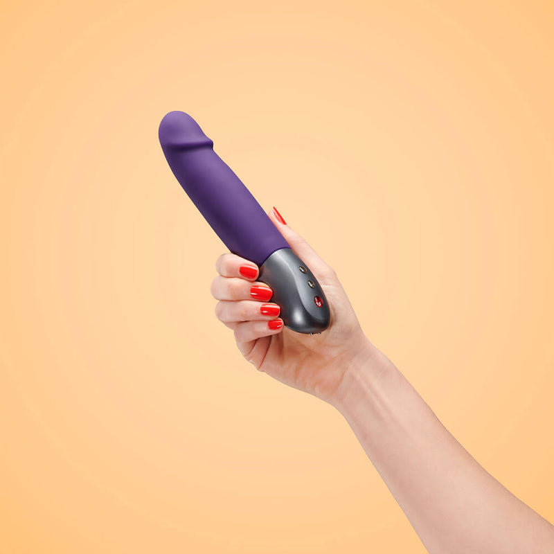 A hand holds the Fun Factory Stronic Real against an orange background. The dildo is clearly longer than the person's hands. The pronounced head is very clearly visible from here. | Kinkly Shop