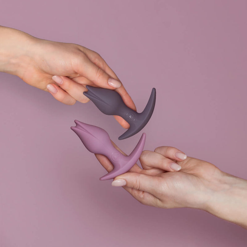 Two hands are holding the two Fun Factory Bootie Fem colors next to one another. Both are shades of purple. The Dark Taupe is a deep shade of purple that looks like purple mixed with black. The Dusk is a lighter shade of purple that looks like it was mixed with a lilac. | Kinkly Shop