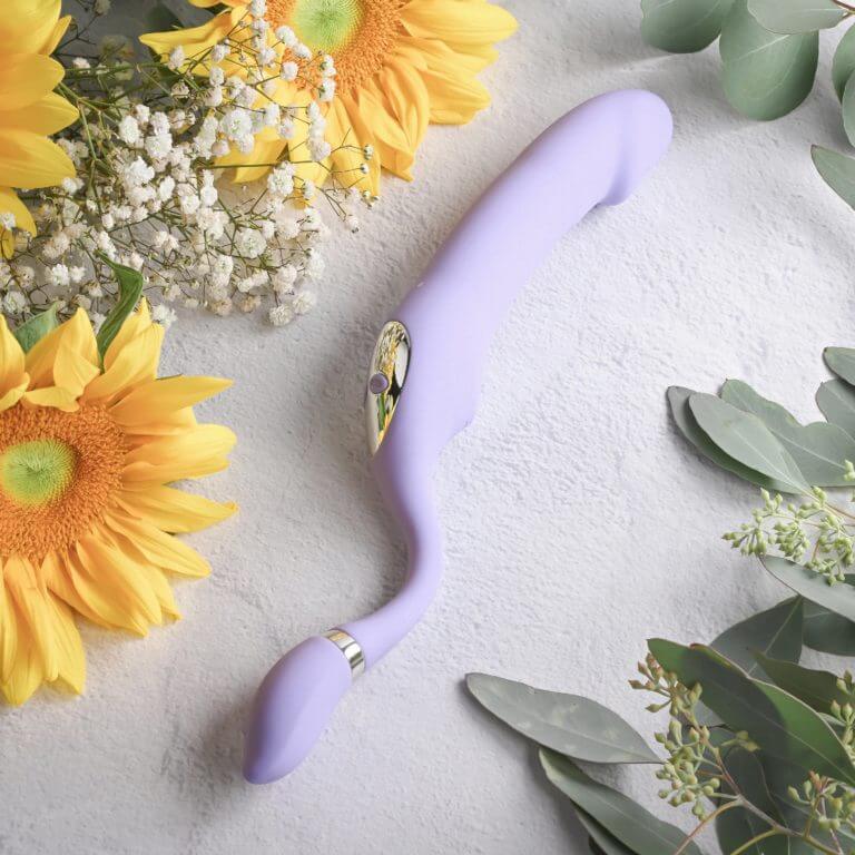 The Gender X Orgasmic Orchid laid flat while surrounded by greenery. The toy looks very long when flattened like this, and the difference in thickness between the smaller vibrating tail and the thicker dildo-like shaft is very noticeable. | Kinkly Shop